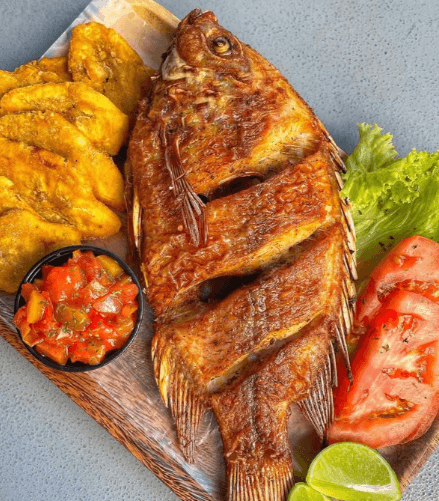 Pescado entero frito y crujiente, acompañado de tostones dorados, ensalada fresca de tomate y lechuga, pico de gallo casero y rodajas de limón, servido sobre una tabla de madera.