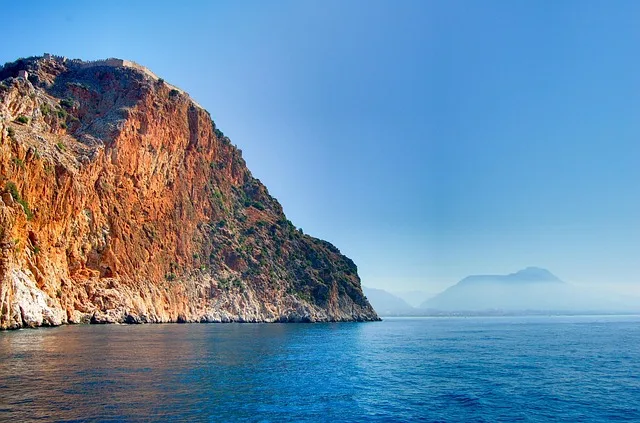 En la imagen una costa marítima con un acantilado que destaca, en el fondo se muestran montañas y el agua es azul como el cielo.