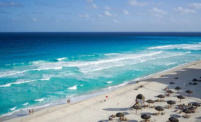 En la imagen una península con aguas con distintos tonos de azul que se mezclan y son armoniosos, se puede ver también arena dorada en la que hay palapas aparentemente vacías.