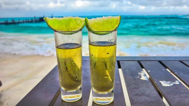 En la imagen dos shots de tequila con un limón con sal en la parte superior, estos sobre una mesa de madera y como fondo esta el mar.