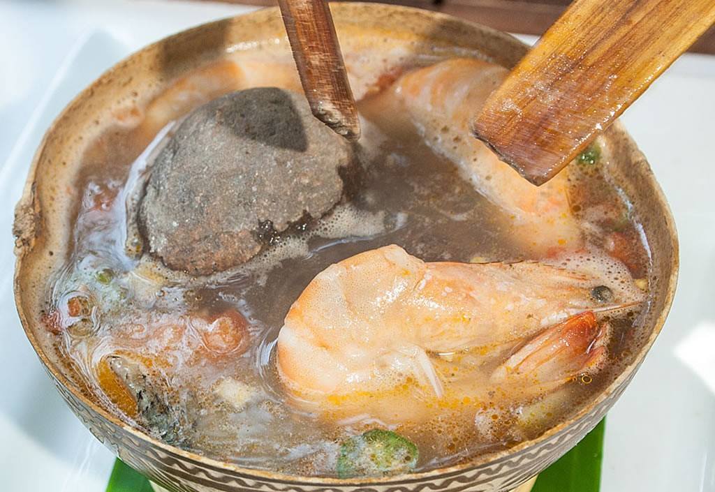 Delicioso caldo de piedra servido en un tazón de madera acompañado de cilantro fresco y chiles, excelente para comer en la playa