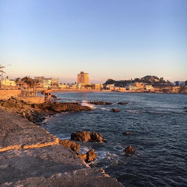 Vista costera al atardecer con edificios iluminados por la luz dorada y olas rompiendo contra las rocas.