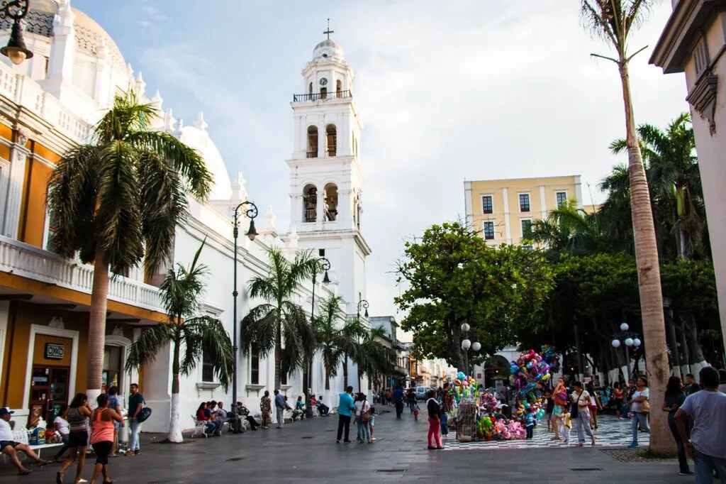 En la imagen un pueblo cercano a una de las playas más bonitas y baratas de México. De fondo una iglesia de color blanca que complementa bien el cielo azul y despejado. Al rededor de la iglesia se ve a personas disfrutando y siendo parte del comercio local.
