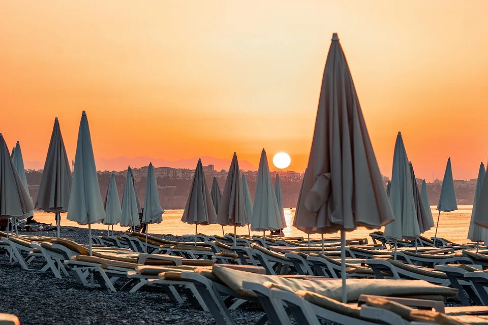 En la imagen se muestra el paisaje de la playa iluminado con el sol en un atardecer con enfoque a a las sombrillas cerradas y camastros vacíos.