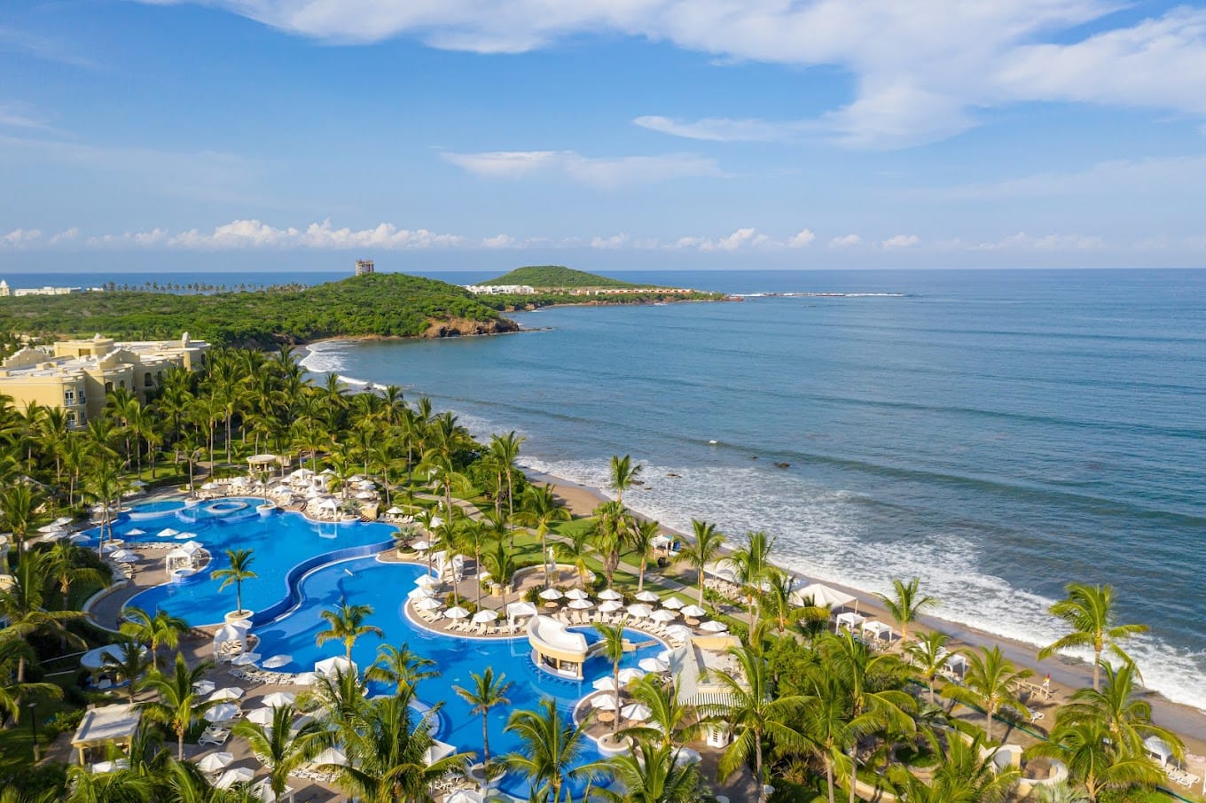 Piscina rodeada de palmeras, frente al mar con vista a las montañas.