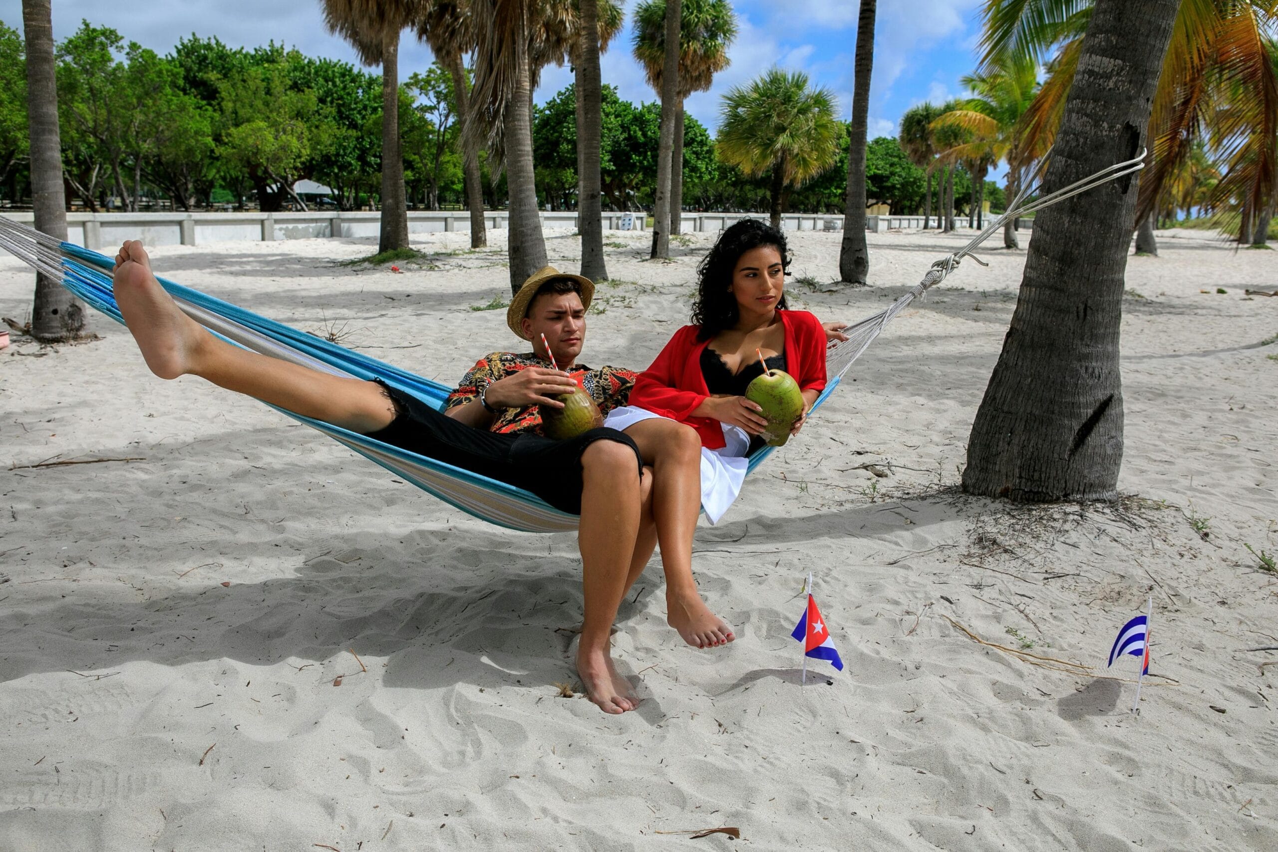 Pareja joven disfrutando de unos cocos y recostados sobre una hamaca azul en la playa de arena blanca durante sus vacaciones, ideal para hablar sobre qué actividades pueden hacer en la playa y rodeados de muchas palmeras a su alrededor.