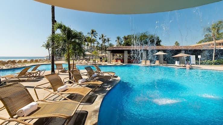 Una alberca con sombra frente al mar, con una palapa al lado, palmeras y camastros alrededor, creando un ambiente tropical y relajante.